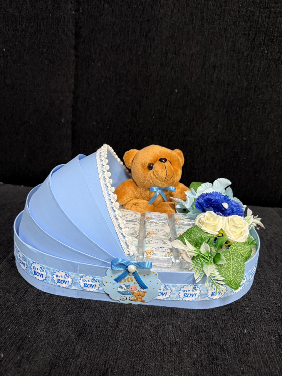 bassinet-shaped gift basket with a teddy bear, foil chocolates, and white and blue flowers, decorated with an "It's a Boy" ribbon for a baby shower gift