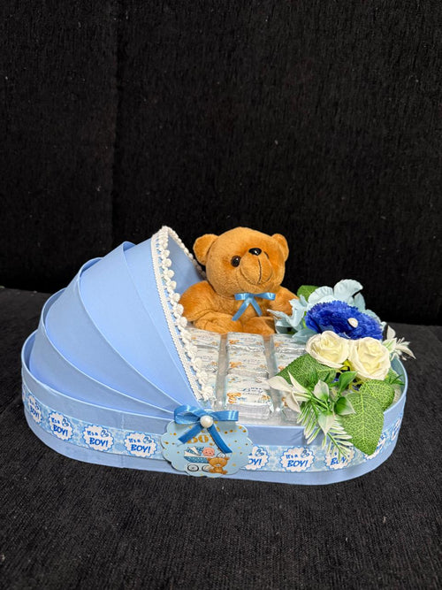 bassinet-shaped gift basket with a teddy bear, foil chocolates, and white and blue flowers, decorated with an "It's a Boy" ribbon for a baby shower gift